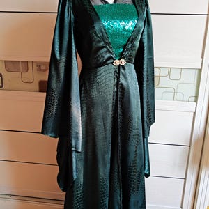 May include: A dark green, floor-length dress with a reptile-like pattern. The dress features long, flowing sleeves and a sequined bodice. A decorative clasp adorns the waist. The dress is displayed on a mannequin.