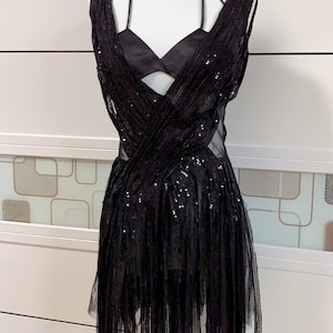 Può includere: Un mini abito nero con paillettes, scollatura profonda e un velo trasparente. L'abito ha un design incrociato sul davanti e un orlo sfrangiato.