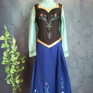Pode incluir: Um traje com um corpete preto com acabamentos dourados e detalhes florais, combinado com uma camisa de manga comprida azul clara e uma saia azul royal. A saia tem uma bainha ondulada azul clara com desenhos florais. O traje é exibido em um manequim.