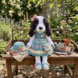 May include: A handcrafted crochet dog doll with a blue dress, matching purse, and sandals. The dog has a pink bow, and a blue hat with floral accents sits nearby. The scene is set on a wooden bench in a garden.