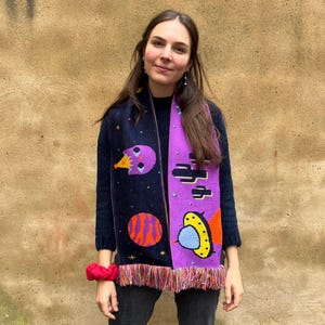 May include: A knitted scarf featuring a space-themed design with planets, stars, and other celestial elements in shades of purple, orange, yellow, and blue. The scarf has a dark blue background and a fringed edge.