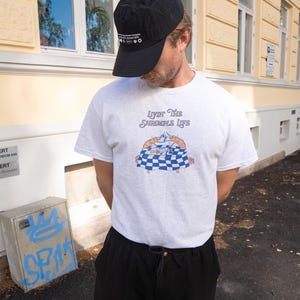 Puede incluir: Gorra de b&eacute;isbol negra y camiseta gris claro con el texto "Livin' The Shrimpole Life" y un gr&aacute;fico de dos camarones sobre un mantel a cuadros. La persona lleva pantalones negros.