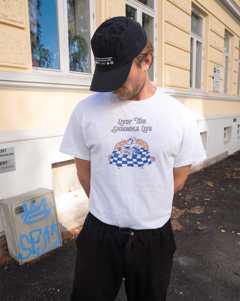 Puede incluir: Gorra de b&eacute;isbol negra y camiseta gris claro con el texto "Livin' The Shrimpole Life" y un gr&aacute;fico de dos camarones sobre un mantel a cuadros. La persona lleva pantalones negros.