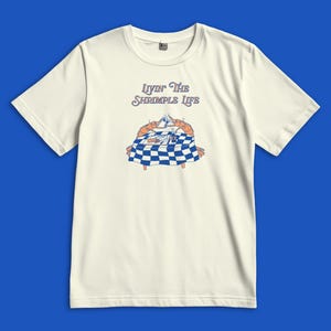 Puede incluir: Una camiseta de manga corta color hueso con un gr&aacute;fico de dos camarones naranjas sentados en una mesa de cuadros azules y blancos, brindando con copas. El texto sobre la ilustraci&oacute;n dice "Livin' The Shrimple Life" en letra azul.