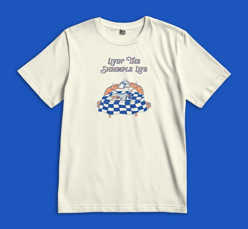 Puede incluir: Una camiseta de manga corta color hueso con un gr&aacute;fico de dos camarones naranjas sentados en una mesa de cuadros azules y blancos, brindando con copas. El texto sobre la ilustraci&oacute;n dice "Livin' The Shrimple Life" en letra azul.