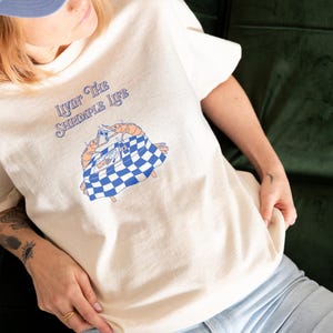 Puede incluir: Una camiseta de color crema con un gr&aacute;fico de dos camarones naranjas sentados a una mesa con un mantel de cuadros azules y blancos. El texto sobre el gr&aacute;fico dice "LIVIN' THE SHRIMPLE LIFE" en letras azules, estilizadas y cursivas.