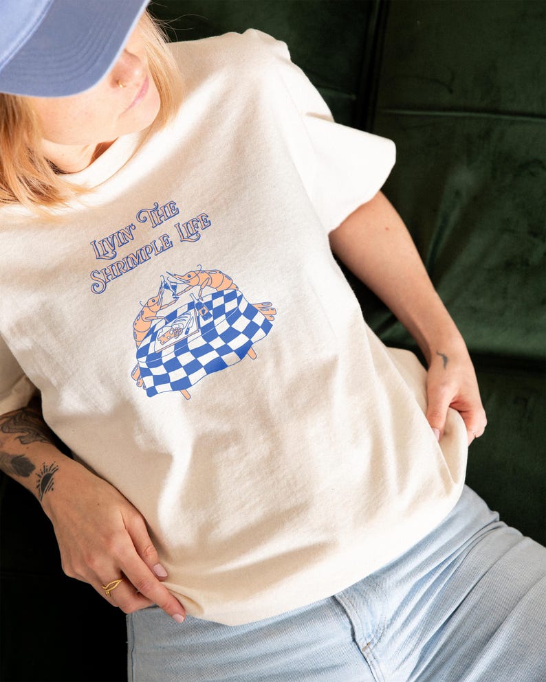 Puede incluir: Una camiseta de color crema con un gr&aacute;fico de dos camarones naranjas sentados a una mesa con un mantel de cuadros azules y blancos. El texto sobre el gr&aacute;fico dice "LIVIN' THE SHRIMPLE LIFE" en letras azules, estilizadas y cursivas.