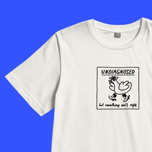 Könnte beinhalten: Ein weißes Kurzarm-T-Shirt mit einer schwarzen Strichzeichnung in einem quadratischen Rahmen. Die Grafik zeigt einen Vogel mit Cowboyhut und Stiefeln, mit dem Text "UNDIAGNOSED" darüber und "but something ain't right" unter dem Vogel.