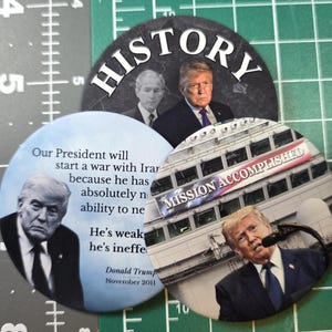 Puede incluir: Un conjunto de tres botones políticos circulares. Un botón presenta la palabra "HISTORY" e imágenes de dos hombres. Otro botón tiene una cita de Donald Trump. El tercer botón muestra a Trump hablando con el texto "MISSION ACCOMPLISHED".