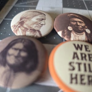 May include: Four round pinback buttons. Three buttons feature sepia-toned portraits of Native American people. The fourth button has the text "WE ARE STILL HERE" in brown letters on a cream background with an orange border.