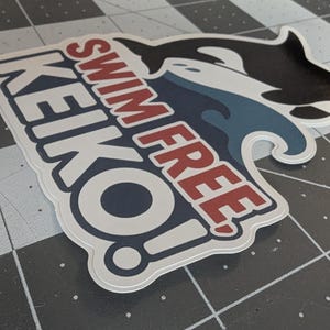 May include: A sticker featuring a black and white orca whale leaping over a blue wave. The sticker has the words "SWIM FREE KEIKO!" in red and white lettering, with a white border.