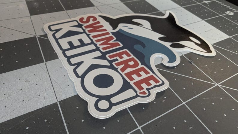May include: A sticker featuring a black and white orca whale leaping over a blue wave. The sticker has the words "SWIM FREE KEIKO!" in red and white lettering, with a white border.