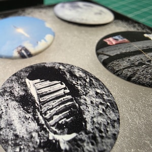 May include: A set of four circular pin-back buttons featuring space-themed imagery, including a black and white lunar boot print, a rocket launch against a blue sky, a view of the moon surface, and an American flag planted on the lunar landscape.