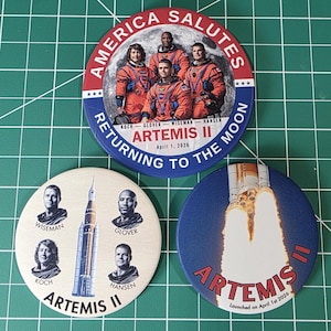 May include: Three round Artemis II themed buttons. One button features the crew in orange suits with the text "America Salutes" and "Returning to the Moon". Another button shows the rocket and the text "Artemis II". The third button displays the crew's portraits.