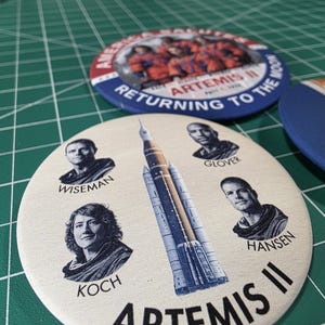 May include: A collection of round buttons featuring the Artemis II mission. One button displays the mission name and crew portraits, while others show the mission logo and text "Returning to the Moon". The buttons are in shades of blue, red, and white.