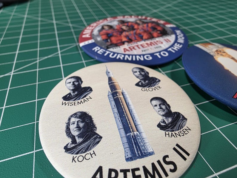 May include: A collection of round buttons featuring the Artemis II mission. One button displays the mission name and crew portraits, while others show the mission logo and text "Returning to the Moon". The buttons are in shades of blue, red, and white.