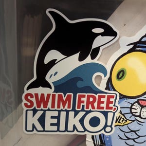 May include: A sticker featuring a black and white orca leaping over a blue wave. The text "SWIM FREE, KEIKO!" is in red and white. The sticker has a white border.