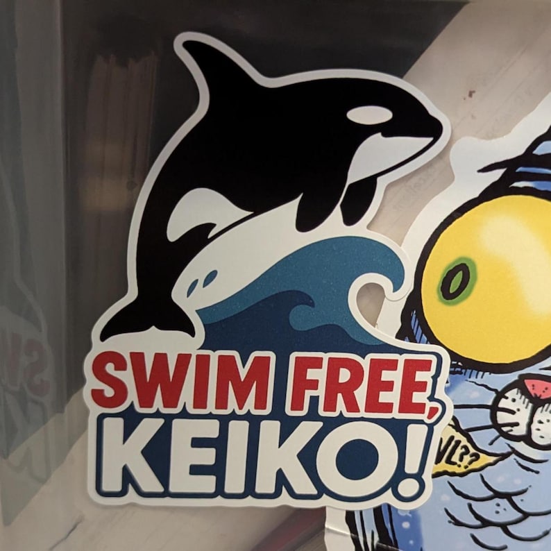 May include: A sticker featuring a black and white orca leaping over a blue wave. The text "SWIM FREE, KEIKO!" is in red and white. The sticker has a white border.
