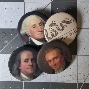 May include: Four round pinback buttons featuring portraits of historical figures and a coiled snake with the text "SIN, or DIE." The buttons depict George Washington, Benjamin Franklin, and other figures. The buttons are black and white.