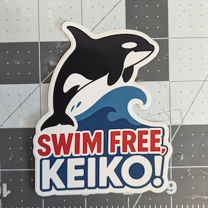 May include: A sticker featuring a black and white orca leaping over a blue and white wave. The text "SWIM FREE, KEIKO!" is in red and white. The sticker is on a gray and black grid.