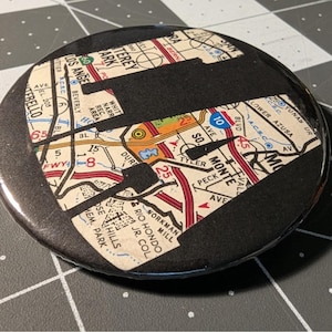 Puede incluir: Botón redondo con un diseño de mapa que muestra Los Ángeles y sus alrededores. El botón tiene un borde negro y un acabado brillante, con una forma negra que cubre parte del mapa.