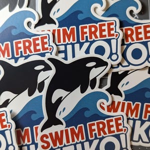 May include: A collection of stickers featuring a black and white orca leaping over blue waves. The stickers have the text "SWIM FREE, KEIKO!" in red and white. The stickers are on a gray cutting mat.