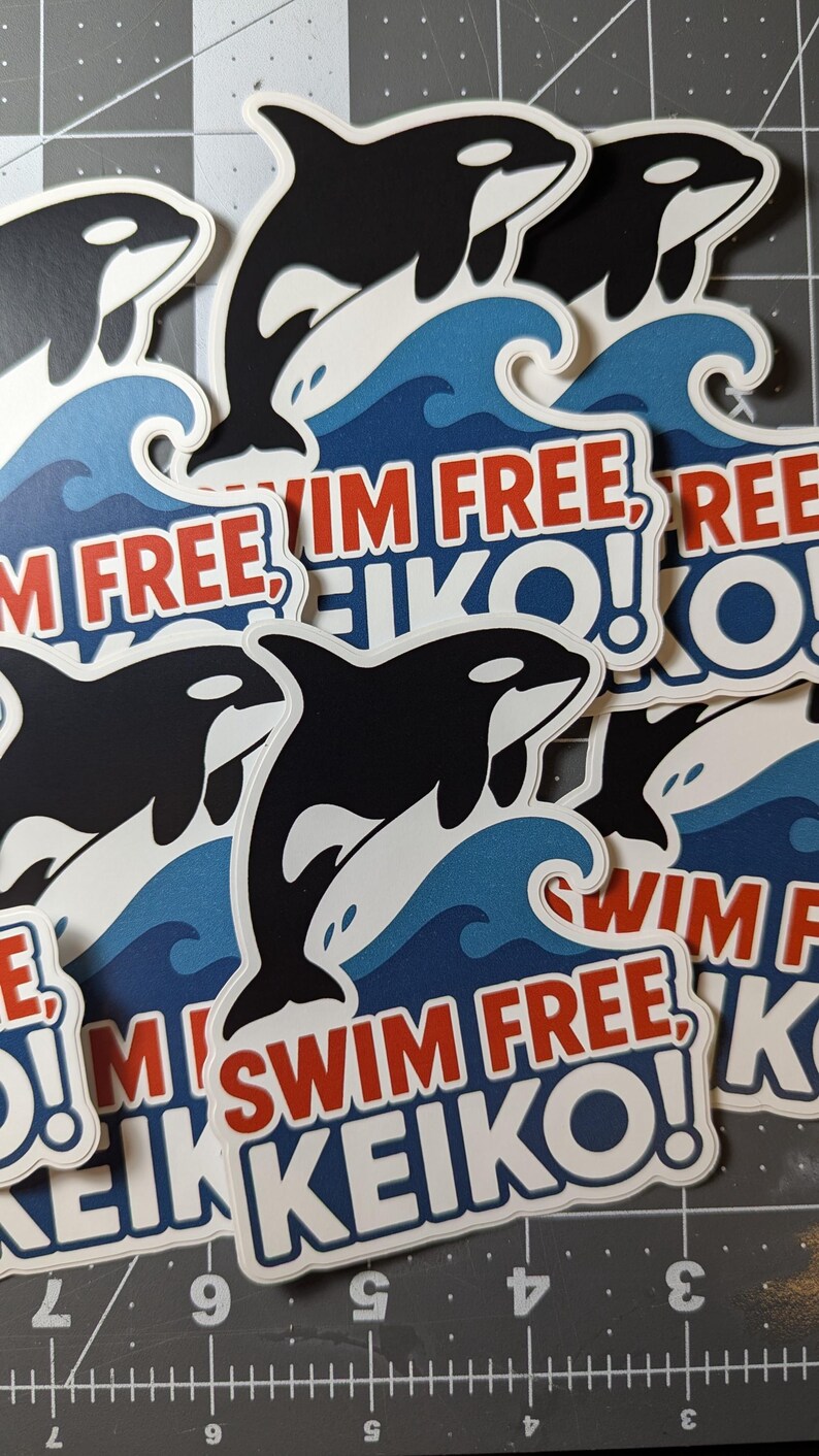 May include: A collection of stickers featuring a black and white orca leaping over blue waves. The stickers have the text "SWIM FREE, KEIKO!" in red and white. The stickers are on a gray cutting mat.