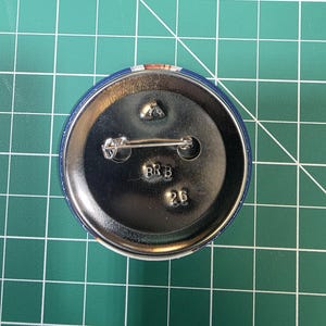 May include: A round, blue button with a metal back and pin. The back of the button has the letters "RRB" and the number "26" stamped on it. The button is on a green grid surface.