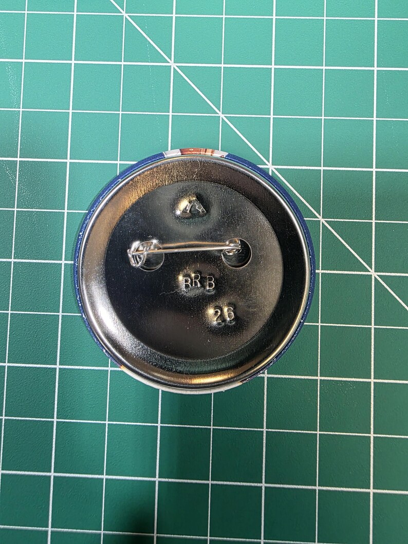 May include: A round, blue button with a metal back and pin. The back of the button has the letters "RRB" and the number "26" stamped on it. The button is on a green grid surface.