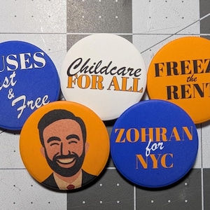 May include: A collection of five round buttons with various designs and text. The buttons are blue, white, and orange. Text includes "Buses Fast & Free", "Childcare For All", "Freeze the Rent", a portrait, and "Zohran for NYC".
