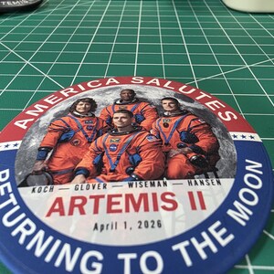May include: A round button with a blue border and the text "RETURNING TO THE MOON." The button features an image of four astronauts in orange suits, with the text "AMERICA SALUTES" and "ARTEMIS II April 1, 2026."