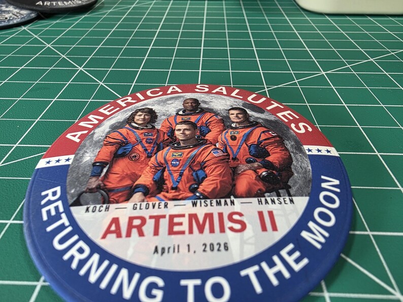 May include: A round button with a blue border and the text "RETURNING TO THE MOON." The button features an image of four astronauts in orange suits, with the text "AMERICA SALUTES" and "ARTEMIS II April 1, 2026."