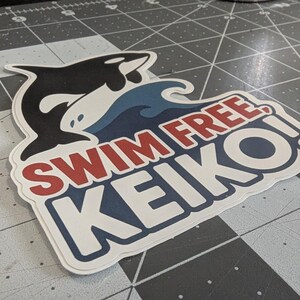 May include: A sticker featuring a black and white orca leaping over a blue wave. The sticker has the text "SWIM FREE KEIKO!" in red and white, with a blue border. The sticker is on a gray cutting mat.