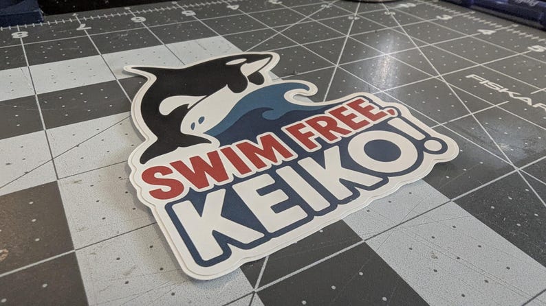 May include: A sticker featuring a black and white orca leaping over a blue wave. The sticker has the text "SWIM FREE KEIKO!" in red and white, with a blue border. The sticker is on a gray cutting mat.