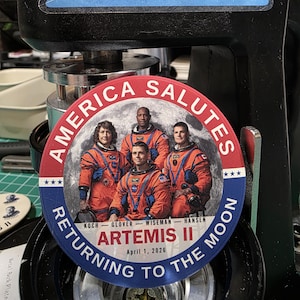 May include: A round badge with the text "AMERICA SALUTES" and "RETURNING TO THE MOON". The badge features a photo of four astronauts in orange suits in front of a moon backdrop. The text "ARTEMIS II April 1, 2026" is also included.