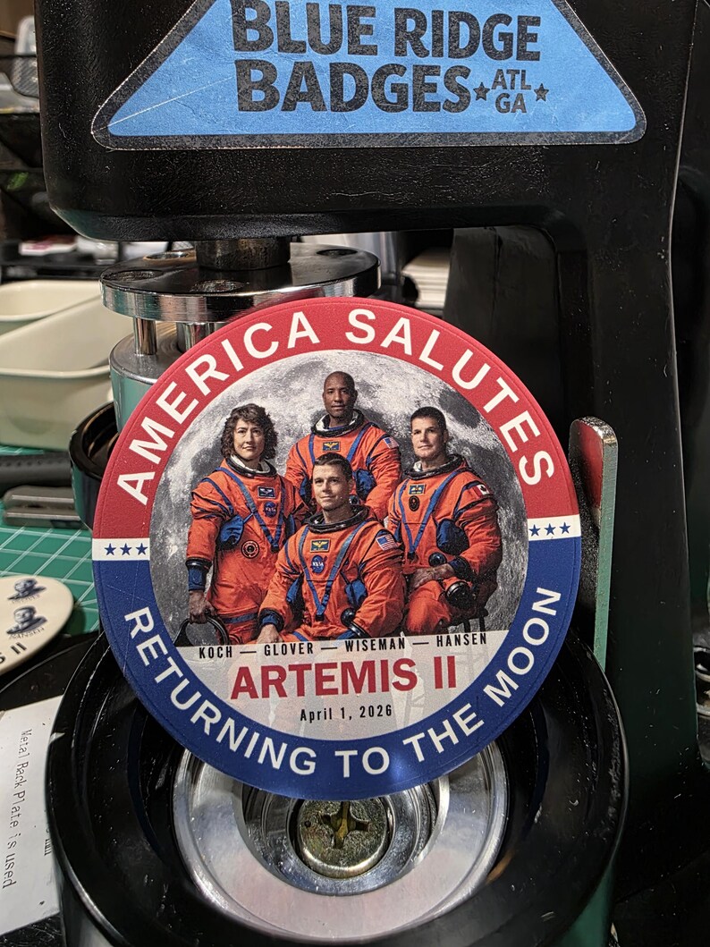 May include: A round badge with the text "AMERICA SALUTES" and "RETURNING TO THE MOON". The badge features a photo of four astronauts in orange suits in front of a moon backdrop. The text "ARTEMIS II April 1, 2026" is also included.