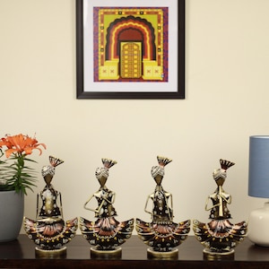 May include: Set of four gold and brown metal figurines of people playing musical instruments. Each figurine is wearing a colourful turban and a long skirt with intricate details. The figurines are standing on a wooden surface.