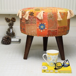 May include: A round, multi-colored patchwork ottoman with wooden legs. The ottoman has a variety of colors and patterns, including orange, yellow, and brown. The ottoman is sitting on a white surface with a book and a cup on top of it.