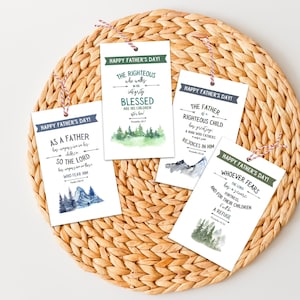 May include: Five rectangular Father's Day gift tags with red and white twine. Each tag features a different inspirational quote and watercolor illustrations of mountains and trees. The tags are arranged on a woven, circular placemat.