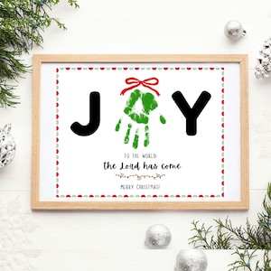 May include: A framed Christmas print with the word "JOY" in black letters. The "O" is a green handprint with a red bow. The text "TO THE WORLD the Lord has come" and "MERRY CHRISTMAS!" are below the word "JOY".