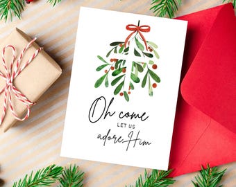 Peut inclure: Carte de Noël blanche avec le texte « Oh come let us adore Him » et un motif de gui à l'aquarelle. La carte est à côté d'une enveloppe rouge, d'un cadeau emballé et de branches de conifères.