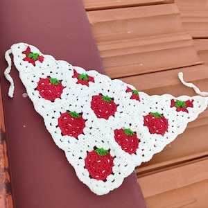 May include: A white crochet bandana featuring a repeating pattern of red strawberries with green leaves. The bandana is triangular in shape with a scalloped edge and ties at the ends. The handmade accessory is displayed on a brown surface.