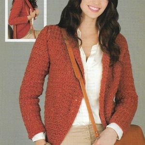 May include: A rust-coloured crocheted cardigan with a shawl collar and long sleeves. The cardigan is open at the front and is being worn over a white button-down shirt and beige trousers.