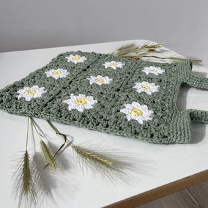 Peut inclure: Un sac cabas en crochet vert avec des fleurs de marguerite blanches. Le sac a deux anses et est pos&eacute; sur une surface blanche.