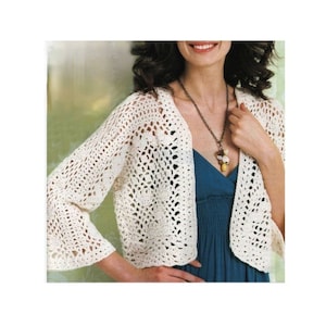 Pode incluir: Bolero de crochê creme com frente aberta e mangas sino. O design leve e rendado apresenta um padrão floral. O suéter estilo bolero é usado sobre um vestido azul e um colar longo.