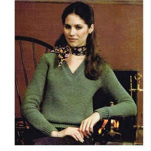 Può includere: Un maglione di lana verde con scollo a V e maniche lunghe. Il maglione è indossato da una donna con lunghi capelli castani e una sciarpa floreale.