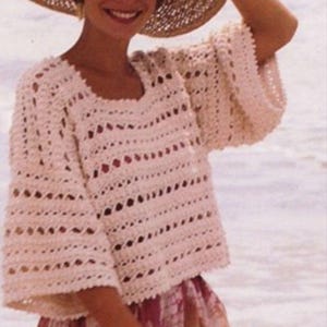 May include: A light beige, open-weave crochet top with wide sleeves and a square neckline. The top is paired with a wide-brimmed straw hat and a patterned skirt. The outfit is suitable for warm weather and beach outings.