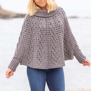 May include: A gray crocheted poncho with a high neck and long sleeves. The poncho has a cable knit pattern and is worn over a pair of blue jeans.