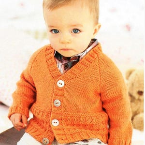 May include: A small child wearing a bright orange knitted cardigan with button closures. The cardigan has a V-neck and is made with a textured stitch.