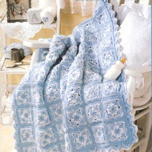 Puede incluir: Una manta de bebé de ganchillo azul claro y blanco con un patrón floral, sobre una silla blanca. La manta tiene un borde festoneado. El texto dice "For Granny's Boy". Un biberón está en la silla.